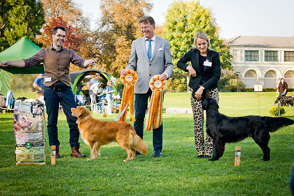 Edison posiert fürs Siegerfoto neben dem Zweitplatzierten, dem Flat Coated Retriever, dahinter stehen die Besitzer und der Richter