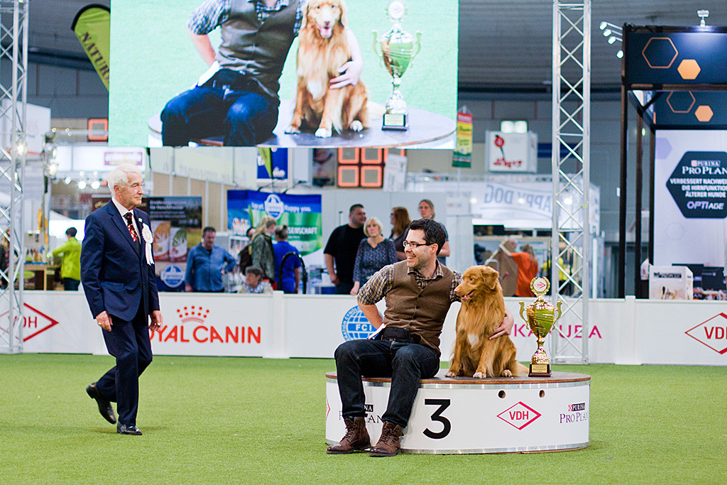 Edison sitzt zusammen mit Stephan auf dem Podest für den dritten Platz, im Hintergrund sind beide auf der großen Leinwand zu sehen, Richter Erwin Deutscher läuft auf sie zu