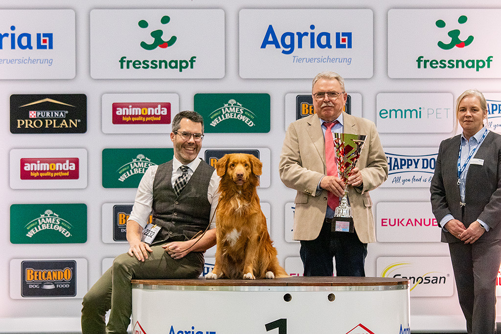 Newton sitzt neben Stephan auf dem Treppchen für Platz 1, dahinter stehend Richter Peter Machetanz, der den Siegerpokal in Händen hält