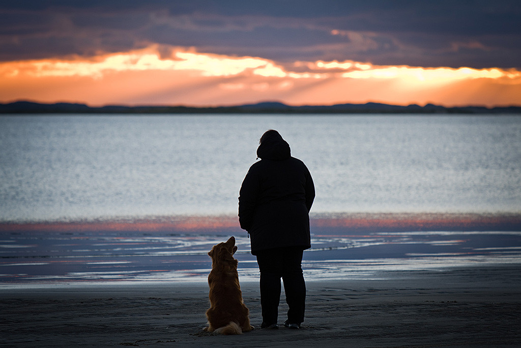 Edison schaut sitzend am Strand von Rømø zu Julia hoch, die gen Meer zum Sonnenuntergang blickt